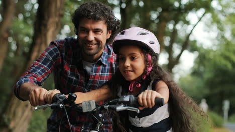 Closeup.-Portrait-of-a-little-girl.-Her-dad-is-teaching-her-to-ride-a-bike.-He-lets-her-go.-Happy-girl.-Moving-camera.-Blurred-background